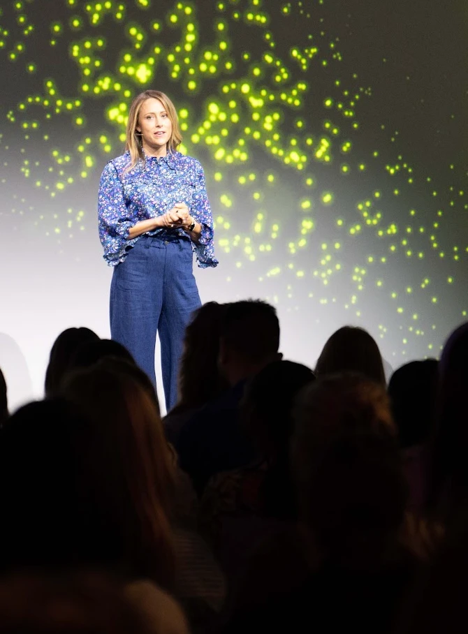 Holly on stage in front of an audience, speaking under a wall of projected lights.