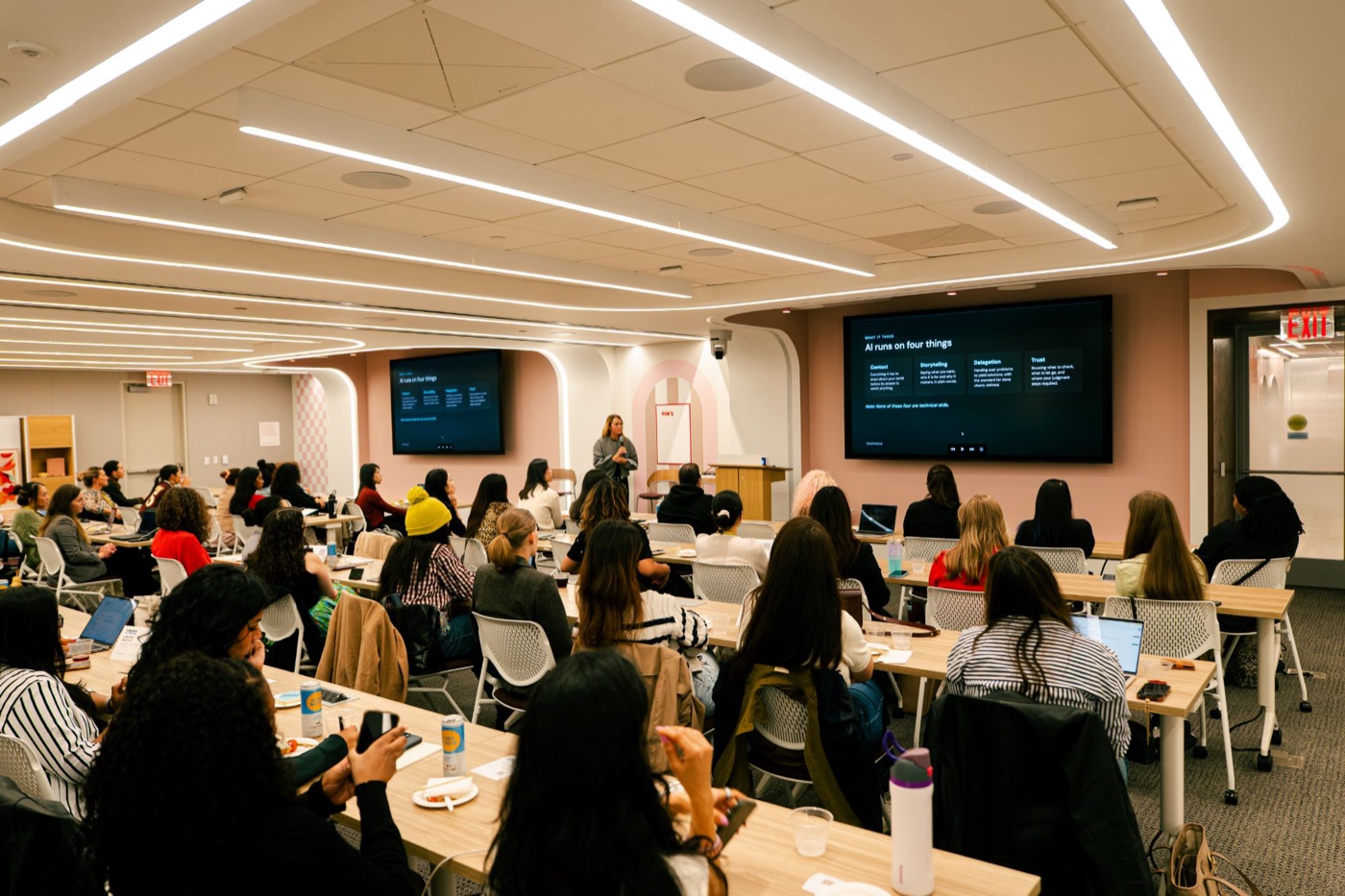 A full room of women at tables, laptops open, facing a screen that reads: AI runs on four things.