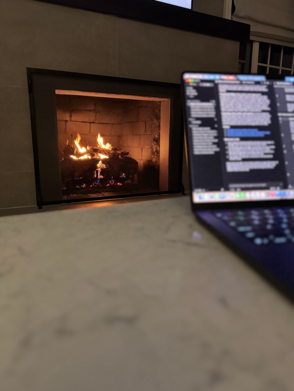 A laptop open on a marble counter before dawn, a fire burning in the fireplace behind it.